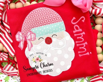 May include: Red Christmas shirt with a white Santa face applique with pink and blue accents. The shirt is personalized with the name "Sammy!" in pink thread. The shirt also has the text "Sawyers Charm Embroidery Designs Est. 2021 Dothan, AL" in black thread.
