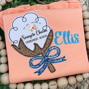 May include: A peach-colored t-shirt with an embroidered cotton boll design. The boll has a brown stem and a blue bow. The design includes the text "Sawyers Charm Embroidery Designs" and "Ellis".
