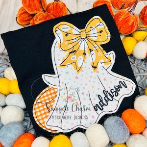 May include: Black t-shirt with a ghost design. The ghost has a yellow bow with the words "Spooky" and "Boo". The ghost's body is white with colorful stars. The name "Addison" is written on the ghost. The design includes the text "Sawyers Charm Embroidery Designs".