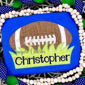 May include: A blue t-shirt with a brown and white football appliqué on a green grass background. The name "Christopher" is embroidered in green on the grass.