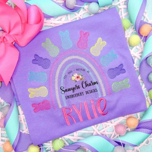 May include: A lilac t-shirt with an embroidered rainbow and colourful bunny shapes. The shirt features the name "Rylie" embroidered in pink. The text "Sawyers Charm Embroidery Designs" is also visible.