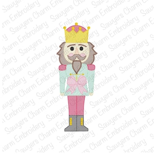 May include: A pink and blue embroidered nutcracker with a gold crown and a pink bow. The nutcracker is standing on a white background.