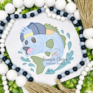 May include: Embroidered design featuring a cartoon fish wearing a baseball cap, with the text "Sawyers Charm Embroidery Designs." The fish is light blue with green and orange accents, and is jumping out of the water. The design is on a white background.