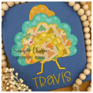 May include: A blue t-shirt featuring an embroidered turkey design. The turkey has a yellow head, a teal body, and a yellow and orange patterned tail. The text "Sawyers Charm Embroidery Designs" and "Travis" is embroidered in yellow on the shirt.