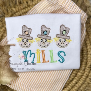 Könnte beinhalten: Ein weißer Strampler mit drei gestickten Vogelscheuchen und dem Namen "MILLS" in bunten Buchstaben. Der Text "Sawyers Charm Embroidery Designs" ist ebenfalls auf den Strampler gestickt.