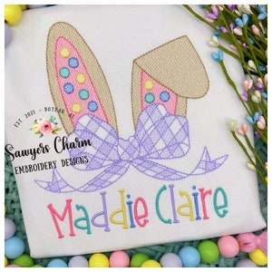 May include: Embroidered design of bunny ears with a purple bow and the name "Maddie Claire" in colorful letters. The design is on a white fabric background.