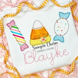 May include: A white t-shirt with a candy corn, two candy pieces, and the name "Blayke" embroidered in pink thread. The text "Sawyers Charm Children's Clothing" is also embroidered on the shirt.