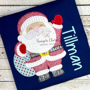 Puede incluir: Camiseta azul marino con un diseño bordado de Papá Noel. Papá Noel está representado con barba blanca, traje rojo y un saco de regalos. El nombre "Tillman" está bordado en azul claro en el lado derecho. El diseño incluye el texto "Sawyers Charms Embroidery Designs".