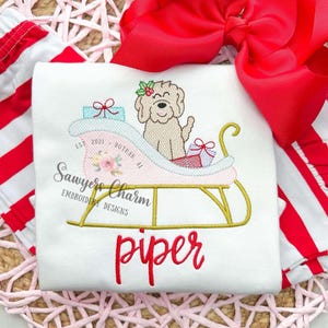 May include: White t-shirt with an embroidered design featuring a dog in a pink sleigh with gifts. The name "piper" is written in red script below the sleigh. The design includes the text "Sawyers Charm Embroidery Designs".