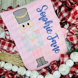May include: A pink embroidered shirt with a nutcracker design and the name "Sophie Jane" embroidered in blue thread.