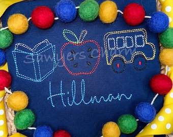 Könnte beinhalten: Ein marineblaues T-Shirt mit einem gestickten Design aus einem Buch, einem Apfel und einem Schulbus. Der Text "Hillman" ist unter dem Design gestickt. Das T-Shirt ist von einer Girlande aus bunten Filzkugeln umgeben.