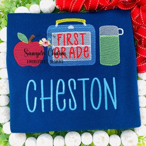May include: A blue t-shirt with embroidered text that says "First Grade" with a red apple, a blue lunchbox, and a green water bottle. The name "Cheston" is embroidered below the text.