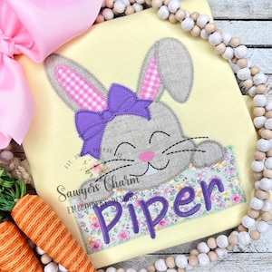 Puede incluir: Una camiseta amarilla con un aplique de conejito sonriente que lleva un lazo morado. El conejito asoma por detrás de un parche floral con el nombre "Piper" bordado en morado.