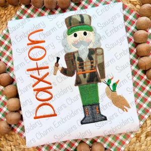 May include: White t-shirt featuring an embroidered nutcracker in camouflage, holding a duck call and two ducks. The name "Doxton" is embroidered in orange. The shirt is on a plaid background.