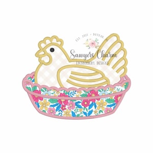 May include: A decorative embroidery design featuring a hen in a floral basket. The hen is white with a gold outline and a black eye. The basket has a pink scalloped edge and is filled with colorful flowers. The text "Sawyers Charm" is also visible.