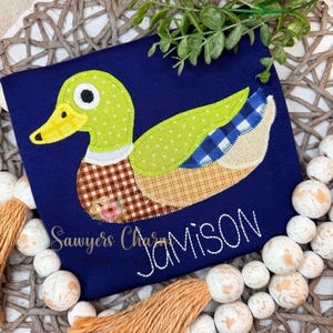 May include: Navy blue shirt with an appliqued duck design. The duck features a yellow beak, green wings, and a patchwork body with plaid and floral patterns. The name "Jamison" is embroidered in white.
