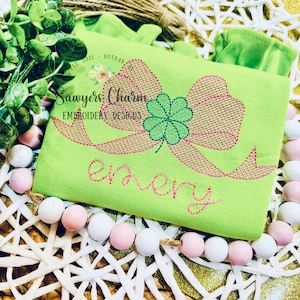 Può includere: Design ricamato verde e marrone di un fiocco con un quadrifoglio su una maglietta verde lime con il testo "Sawyers Charm Embroidery Designs" e il nome "Emery" ricamato in rosa.