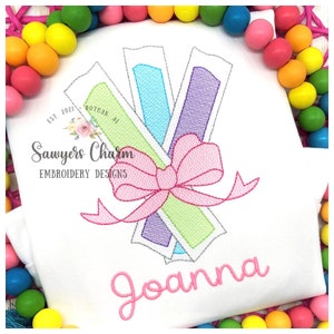 May include: Embroidered design of three colorful pencils tied together with a pink bow. The name "Joanna" is embroidered below the pencils. The design is on a white t-shirt.