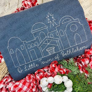May include: White outline embroidery design on a dark blue fabric background. The design depicts a simple outline of the town of Bethlehem with the text "O Little Town of Bethlehem".