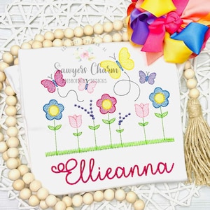 May include: White t-shirt with embroidered flowers, butterflies, and the name "Ellieanna" in pink script. The design includes blue, pink, and yellow flowers, with green stems and grass. A colorful bow is in the upper right corner.