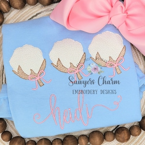 May include: Light blue baby shirt with embroidered cotton bolls and pink bows. The name "Heidi" is embroidered in pink script. A large pink bow is in the background. The text "Sawyers Charm Embroidery Designs" is also visible.