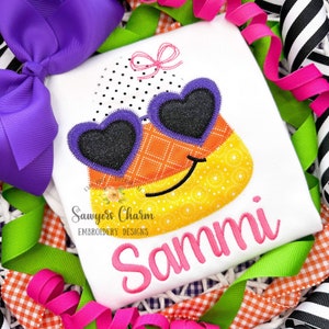 May include: A white t-shirt with an embroidered candy corn design wearing heart-shaped sunglasses. The candy corn has a yellow smile and a pink bow. The text "Sawyers Charm Embroidery Designs" and the name "Sammi" are embroidered on the shirt.