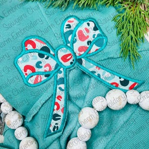 May include: A teal blue fabric with a large, embroidered, red, white and blue bow with a patterned design. The bow is centred on the fabric.