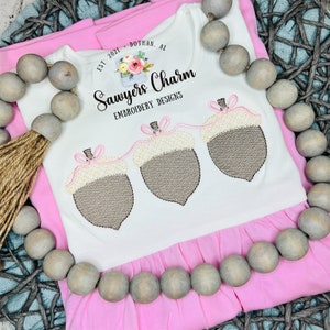 May include: White baby onesie with a pink ruffle trim and a brown embroidered acorn garland. The onesie has the text "Sawyers Charm Embroidery Designs" and "Est. 2021 Dothan, AL" printed on it.