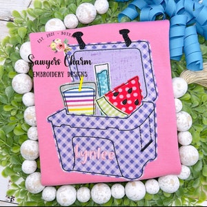 May include: A purple and white checkered cooler with a watermelon, a glass of juice with a straw, and a glass bottle inside. The cooler has a handle and a personalized name "Lynlee" on the front. The text "Sawyers Charm Embroidery Designs" is visible on the pink background.