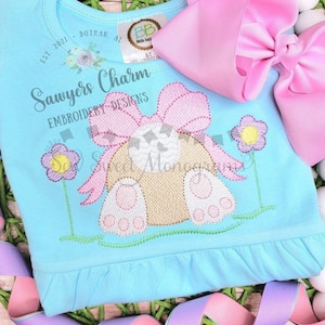 May include: A light blue baby onesie with a white embroidered bunny with a pink bow. The onesie has the text "Sawyers Charm Embroidery Designs" on the front.