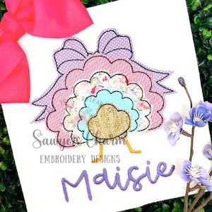 May include: A white t-shirt with a colourful turkey design and a large pink bow. The turkey has layered, patterned feathers in pink, blue, and floral prints. The name "Maisie" is embroidered in purple script below the turkey.