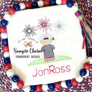 May include: Embroidered design of a child wearing a baseball cap and a blue shirt, standing on a green lawn and looking up at three fireworks in red, white, and blue. The text "Sawyers Charm Embroidery Designs" and "Jon Ross" are also embroidered on the design.