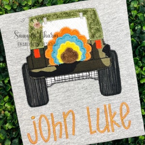 May include: A green camouflage jeep with a flower design on the back. The jeep has a black grill and black tires. The name "John Luke" is embroidered in orange below the jeep.