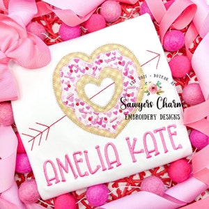 May include: A white t-shirt with pink embroidery of a heart-shaped donut with sprinkles and an arrow through it. The text "Amelia Kate" is embroidered below the donut. The text "Sawyers Charm Embroidery Designs" is embroidered below the heart.