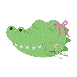 May include: A green embroidered alligator head applique featuring black eyes, a smiling mouth with white teeth, and a pink bow attached to the side. The design includes the text Sawyers Charm Embroidery Designs and Est 2021 with a floral graphic.