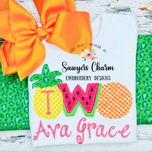 May include: White onesie with embroidered fruit designs spelling out "TWO" and the name "Ava Grace". The onesie is decorated with a yellow bow and a green and black leopard print fabric.