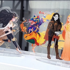 May include: Acrylic standees with vibrant illustrations of fantasy characters. The standees showcase a warrior with a sword, a creature with a tiger, a figure with a sword and cape, and a woman in a gown. The artist's name, "KELSEY ENG," is visible.
