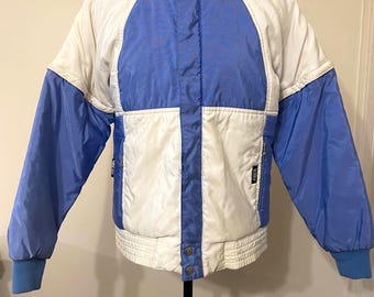 May include: A vintage ski jacket in white and blue. The jacket has a high collar, a zip-up front, and a colour-blocked design with blue sleeves and accents. The jacket has a ribbed hem and cuffs.