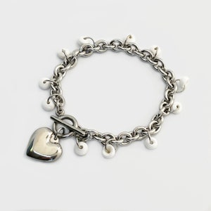 Puede incluir: Un brazalete de cadena de plata con un dije de corazón y cuentas blancas.