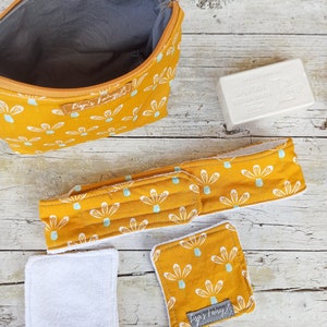Può includere: Una trousse da trucco, una fascia per capelli e due dischetti di cotone con stampa floreale gialla e bianca. La trousse da trucco ha una chiusura a zip e la fascia ha una chiusura a velcro. I dischetti di cotone sono bianchi e hanno una texture morbida. La trousse da trucco ha la scritta "Lizi's Fairy" stampata. La fascia ha la scritta "Lizi's Fairy" stampata. La saponetta è bianca e ha la scritta "Savon Extra Doux" stampata.