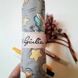 Può includere: Un astuccio per matite in tessuto grigio con una stampa di unicorno, stelle e nuvole. L'astuccio ha una fascia in tessuto rosa con il nome "Giulia" ricamato.