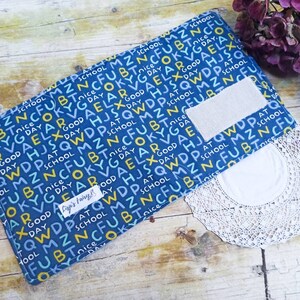 Può includere: Una pochette con cerniera in tessuto blu con una tasca in tessuto bianco. La pochette è ricoperta da un motivo ripetuto di lettere gialle e blu che compongono le parole "scuola", "buona giornata" e "buona giornata".