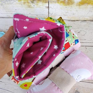 Può includere: Un insieme di articoli in tessuto colorato e fantasia. Tra questi, un elemento arrotolato con interno magenta e motivo a stelle bianche, e un elemento rosa con motivo a nuvole e stelle bianche. Un cinturino beige tiene insieme gli articoli.