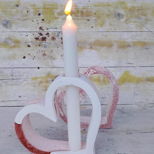 Può includere: Un portacandele bianco a forma di cuore con finiture glitterate rosse, che tiene una candela bianca accesa. La fiamma della candela è arancione brillante. Una seconda decorazione a forma di cuore è dietro il portacandele. Lo sfondo è in legno chiaro.