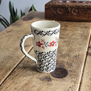 May include: A white ceramic mug with a black and red floral design. The mug has a black floral pattern at the top and bottom, with a red floral band in the middle. The handle is also decorated with the black floral pattern. The mug is on a wooden surface.