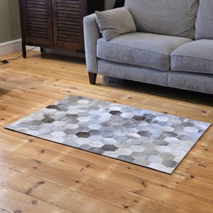 May include: A gray and white cowhide rug with a hexagonal pattern. The rug is laid out on a wooden floor in front of a gray sofa.