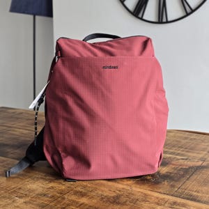 May include: A burgundy backpack with the brand name "mindasa" printed in black. The backpack has a top handle, a front pocket, and a black zip. A white tag hangs from the side. The backpack is on a wooden surface.