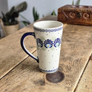 Op de afbeelding: Hoge keramische mok met een witte basis, versierd met blauwe en lichtgroene bloempatronen. De rand en het oor zijn diepblauw. De mok is versierd met kleine blauwe stippen en staat op een houten oppervlak.