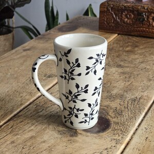 May include: A white ceramic mug featuring a black leaf pattern. The mug has a tall, slender shape with a curved handle. The leaf design is repeated around the mug's exterior. The mug is on a wooden surface.