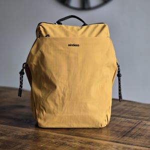 May include: A mustard-yellow backpack with a black handle and zip. The word "mindesa" is printed in black on the front. The bag has black and white rope-like details on the sides. The backpack is on a wooden surface.